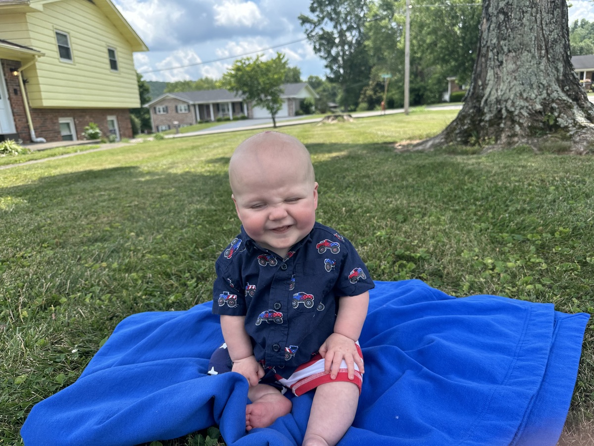 A baby smiles while sitting on a blanket outside in the grass